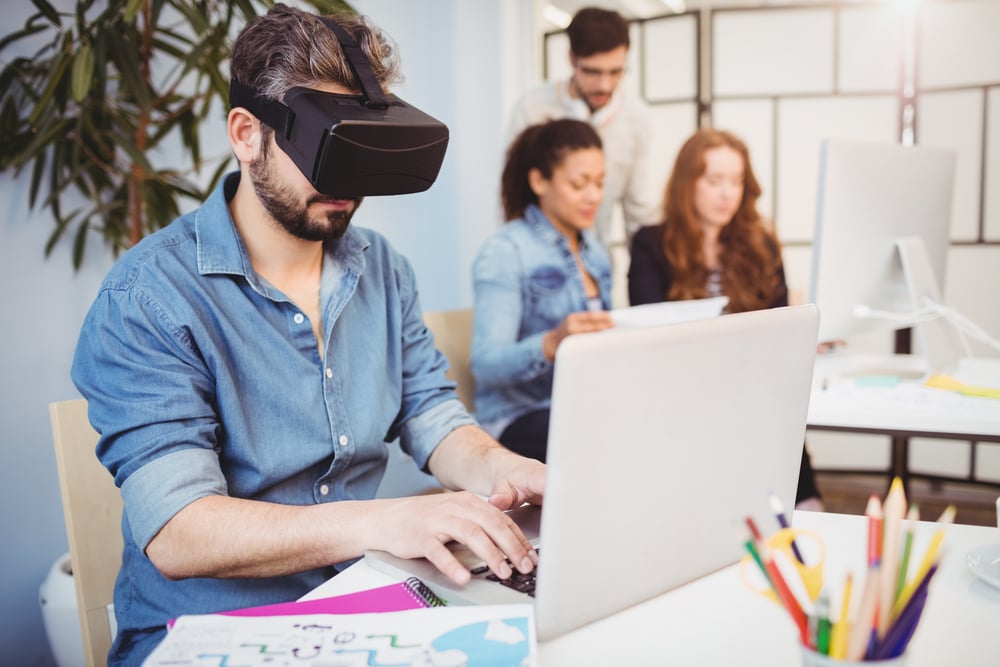 Businessman wearing virtual reality headset while using laptop against colleagues in creative office
