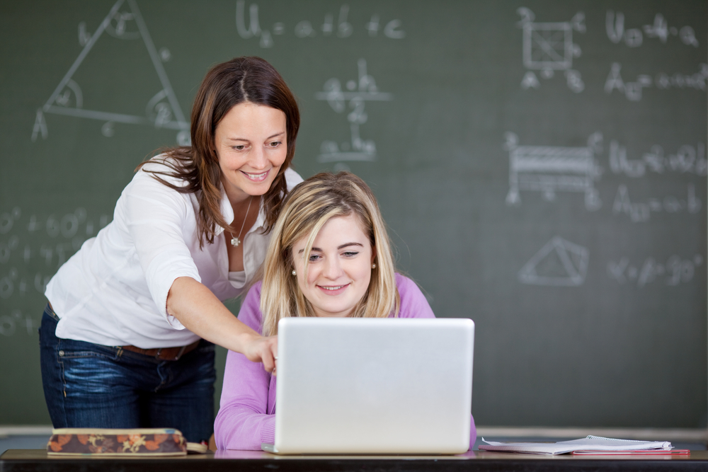 Female teacher assisting student in using laptop at desk in classroom