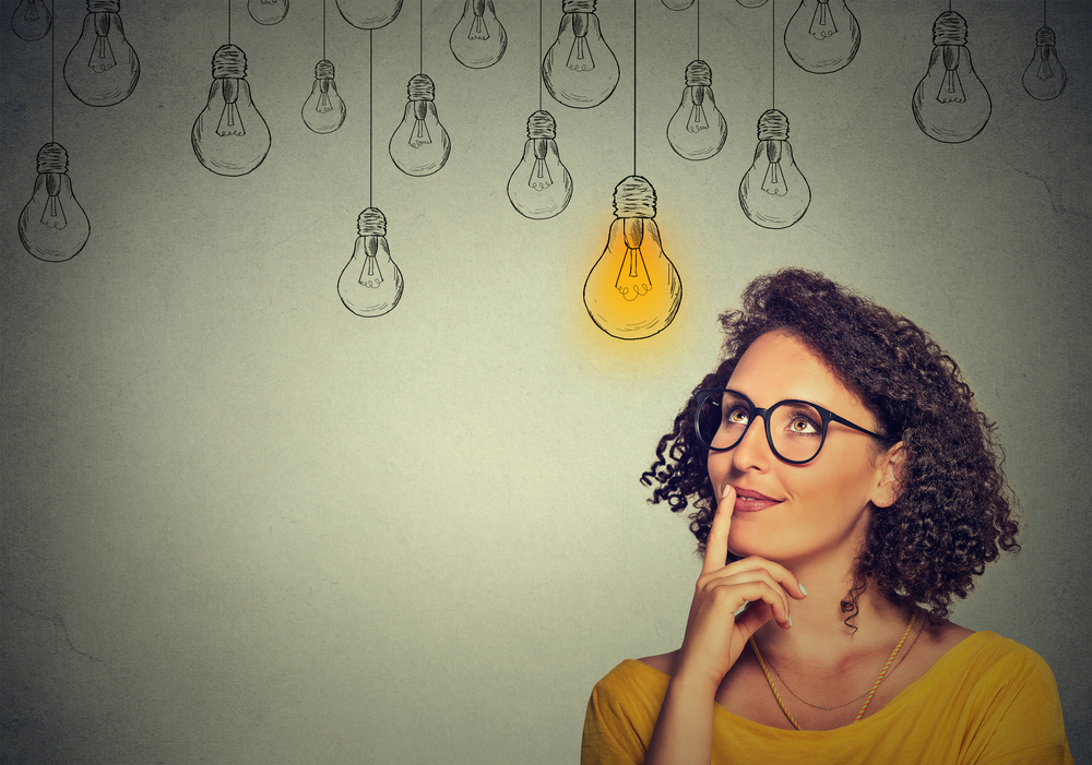 Thinking woman in glasses looking up with light idea bulb above head isolated on gray wall background-2